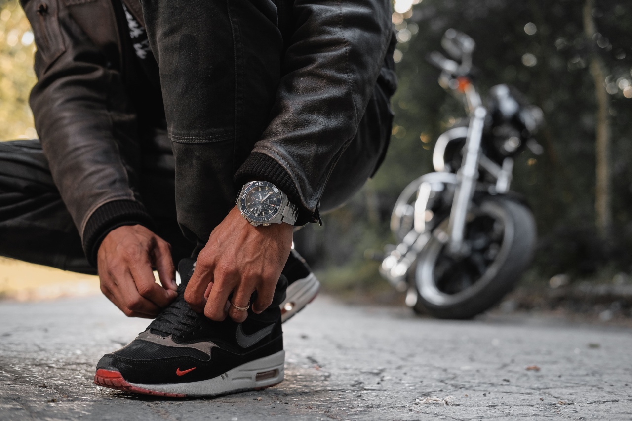 A biker in a leather jacket crouching to tie a shoe with a luxury silver chronograph wristwatch with a dark dial.