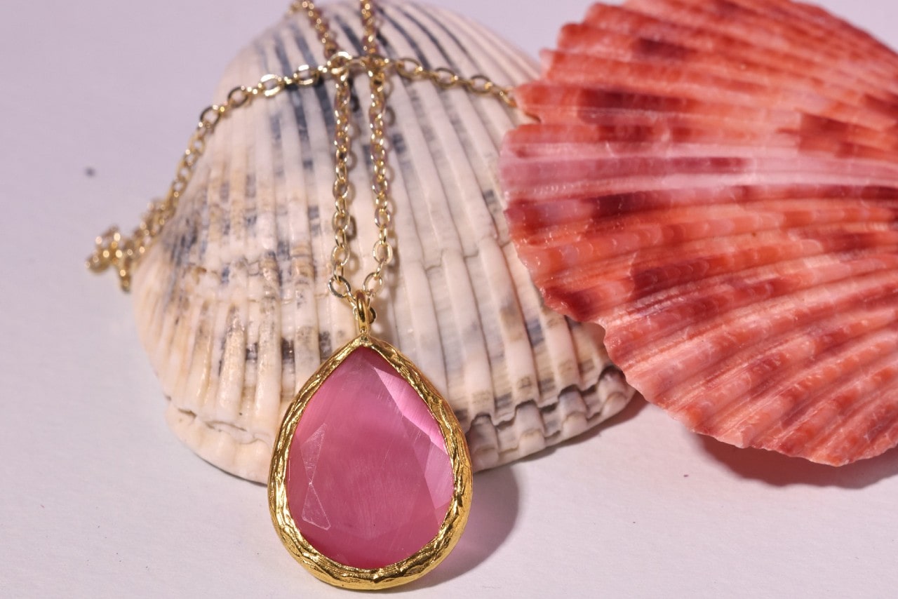 A large pink gemstone pendant with a gold chain resting on shells.