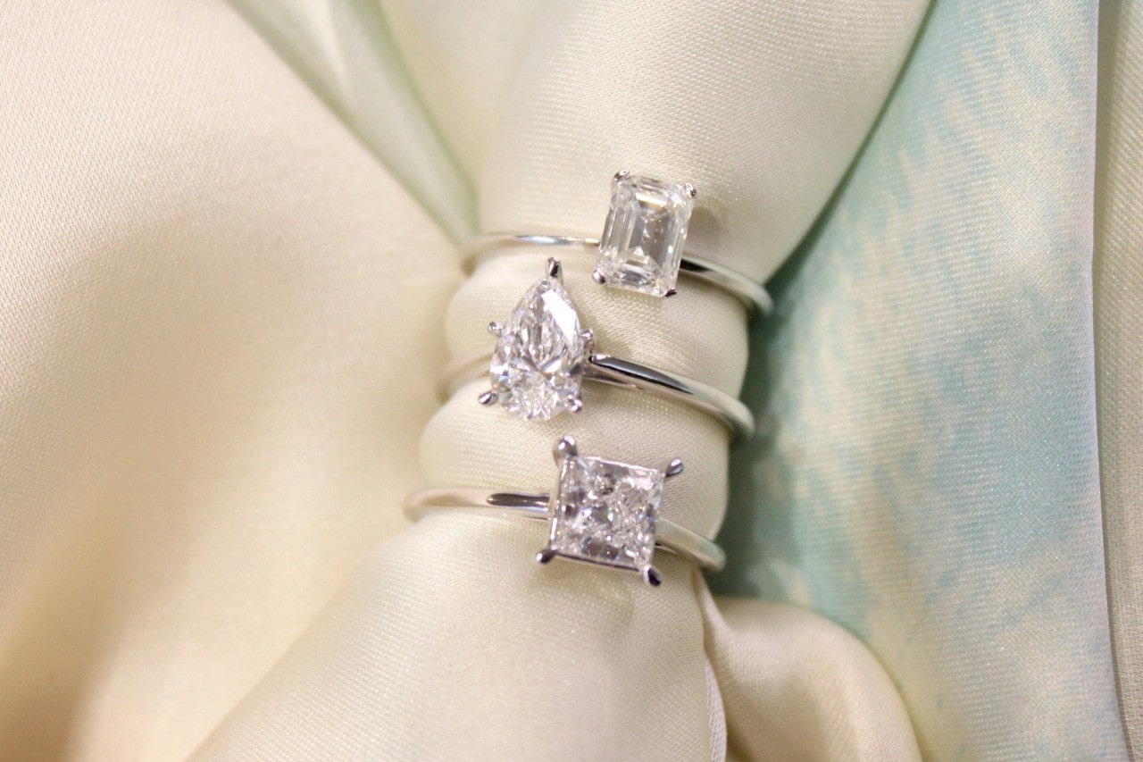 three solitaire engagement rings with different center stone shapes on a white piece of fabric