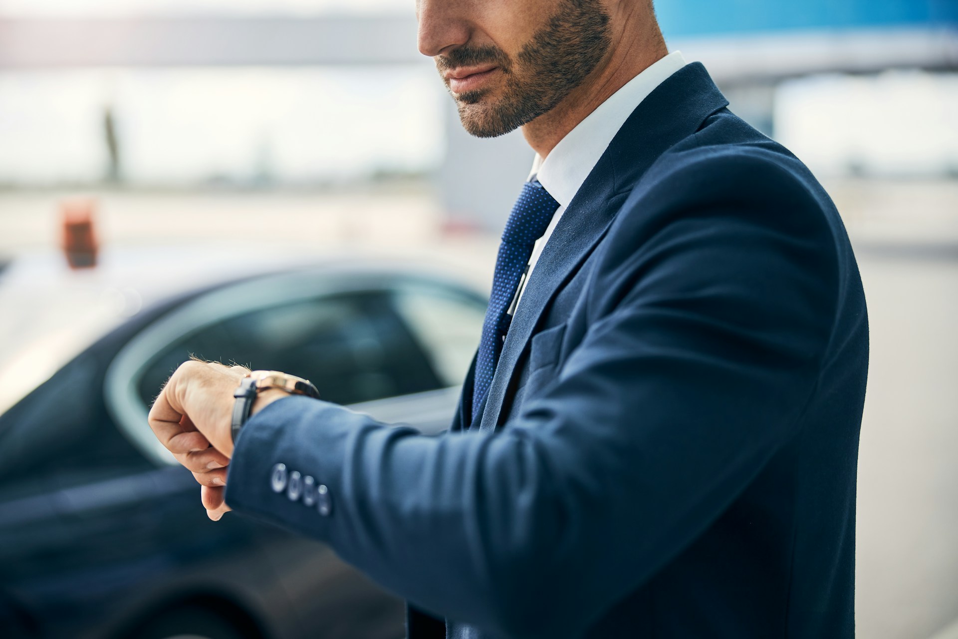 A shot of a well-dressed man in a navy blue suit checking his luxury watch as he walks toward his car.