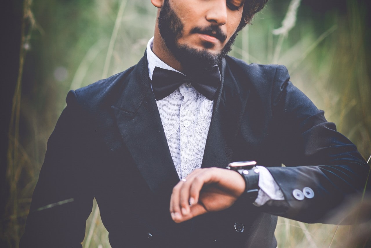 A well-dressed man standing in tall grass, checking his watch.