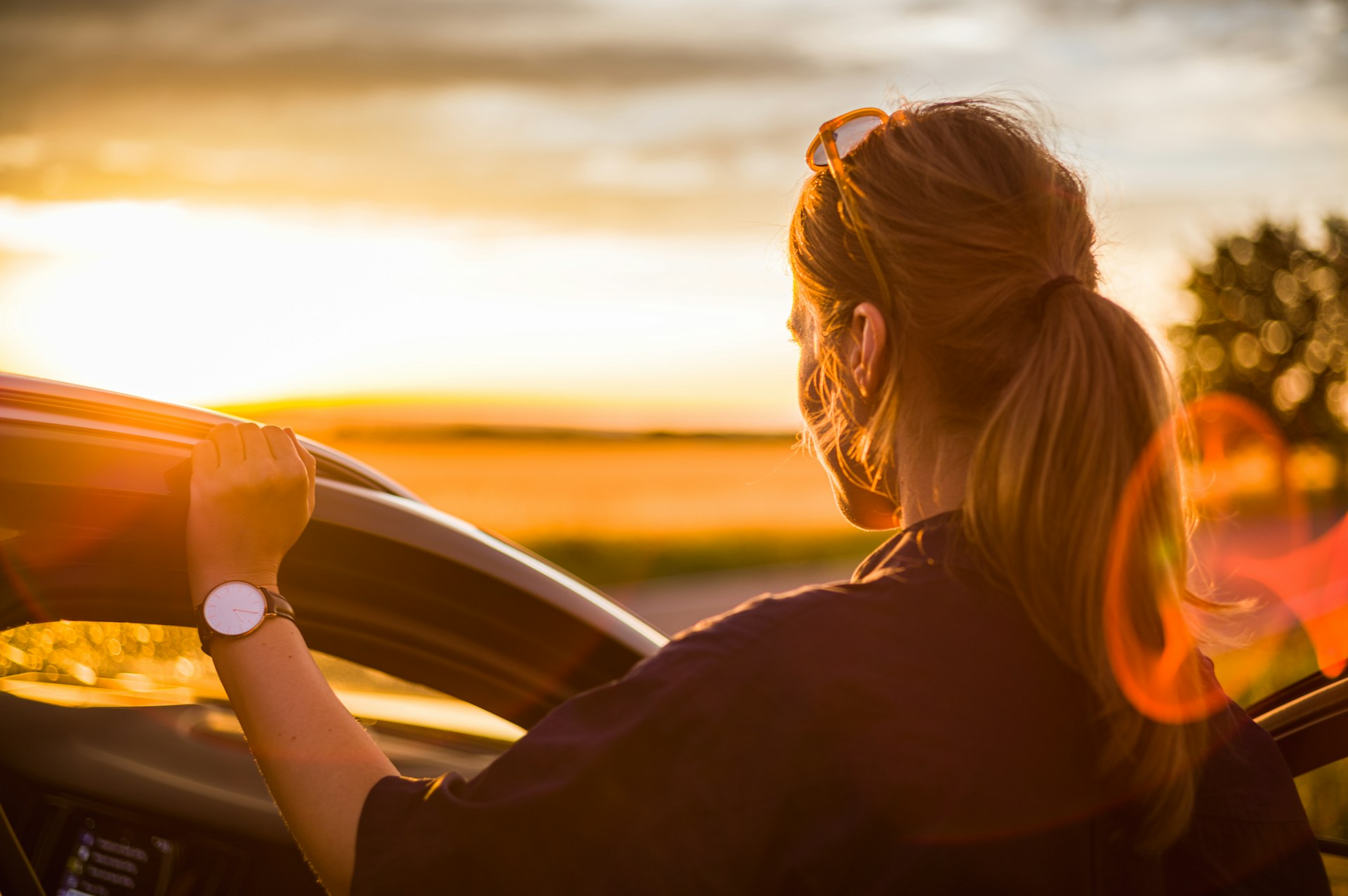 A woman wearing a fine watch resting her hand on her car and looking towards the sunset.